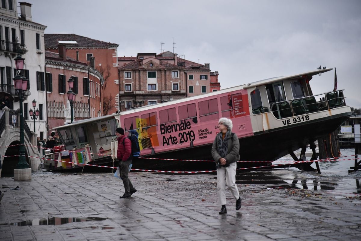 La gente camina junto a un bote de taxis varado en Riva degli Schiavoni, después de este fuera arrastrado durante la marea alta, "Alta Acqua", de la madrugada del 13 de noviembre. (AFP)<br>